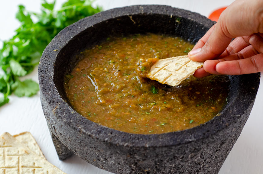 Salsa molcajeteada in a large molcajete surrounded by tomatoes, cilantro, and garlic