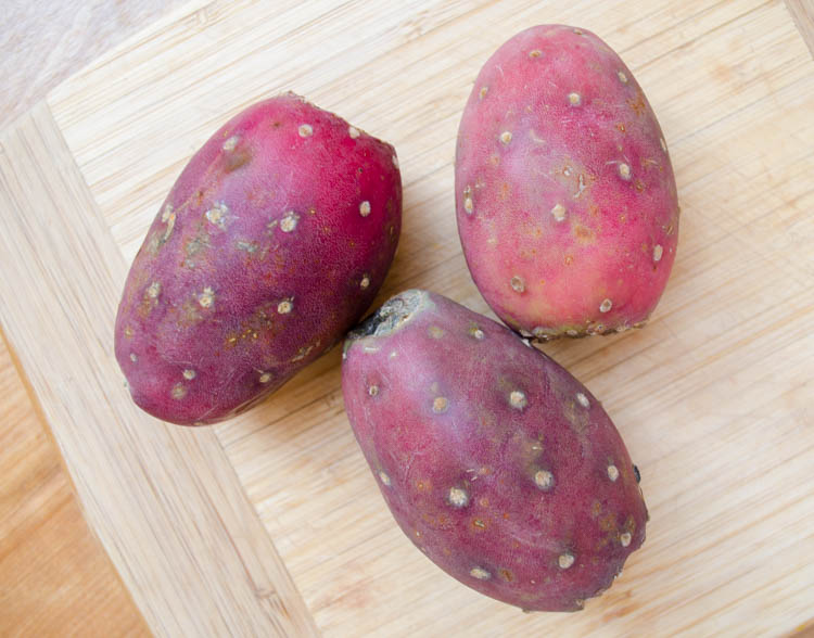 Three fresh red prickly pears on a wood cutting board.