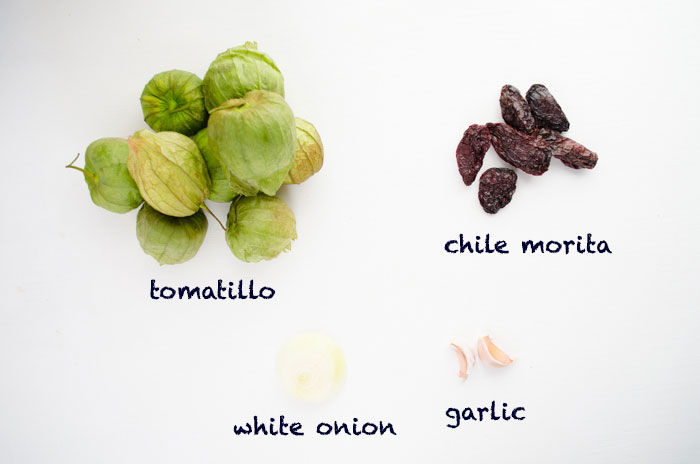 Ingredients on a white surface: tomatillos, chile morita, white onion, and garlic cloves.