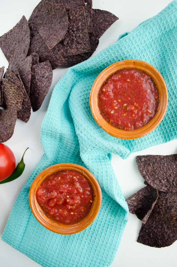 Two small brown bowls of tomato salsa on a teal towel next to blue tortilla chips.