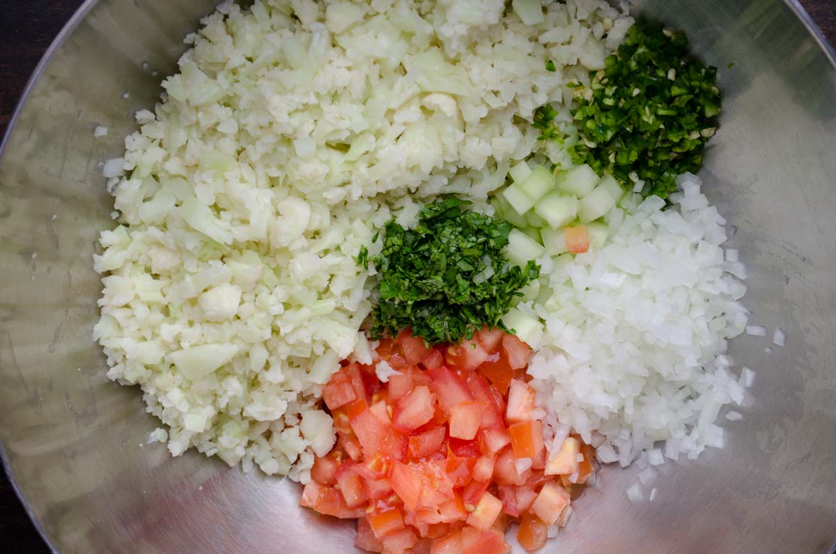 Top view of a metal mixing bowl filled with riced cauliflower, diced tomato and onion, and lots of seasoning.