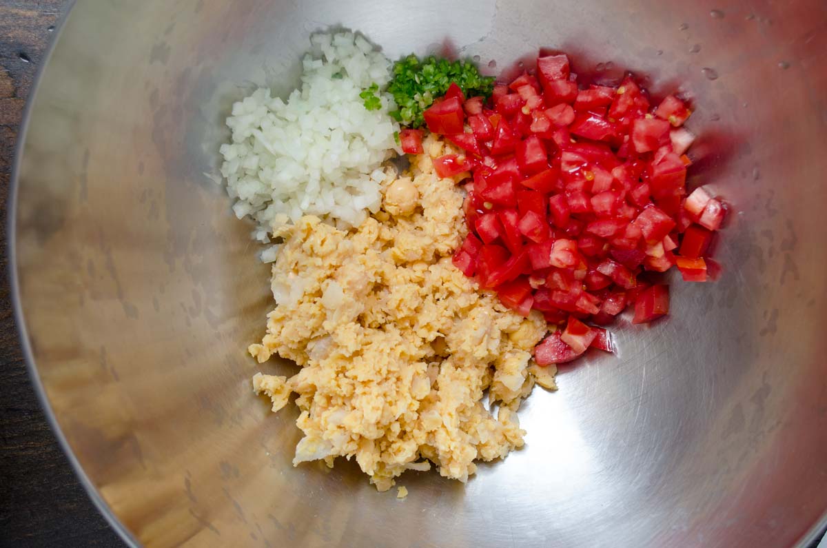 Mashed chickpeas with diced tomato and onion in a metal mixing bowl.