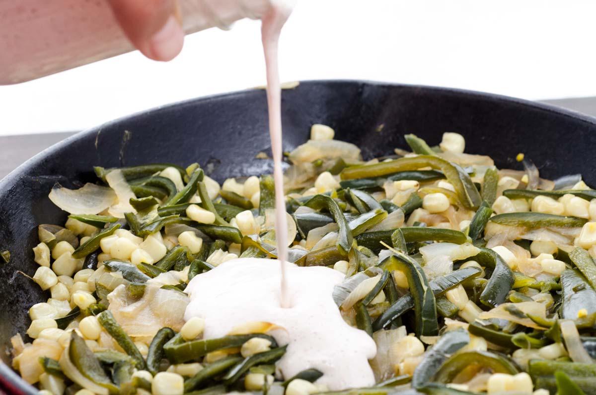 Crema being poured over a cast iron skillet of onions, poblano, and corn.