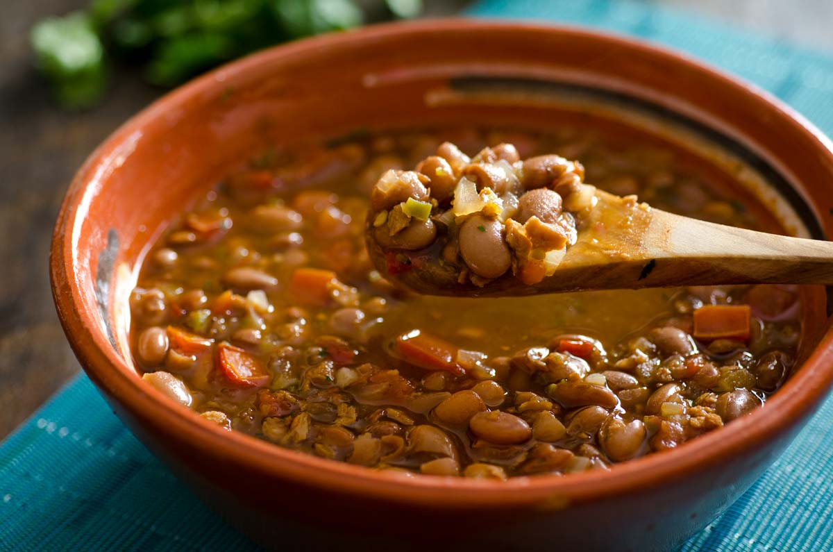 A wooden spoon of Vegan Frijoles Charros over a brown clay bowl.