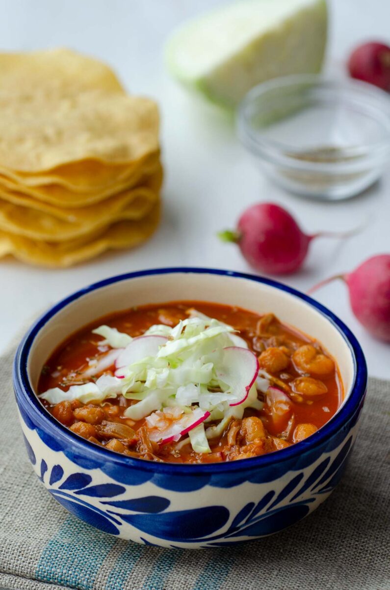 vegan pozole in a white and blue talavera bowl on top of a linen napkin surrounded by radishes, cabbage, and tostadas
