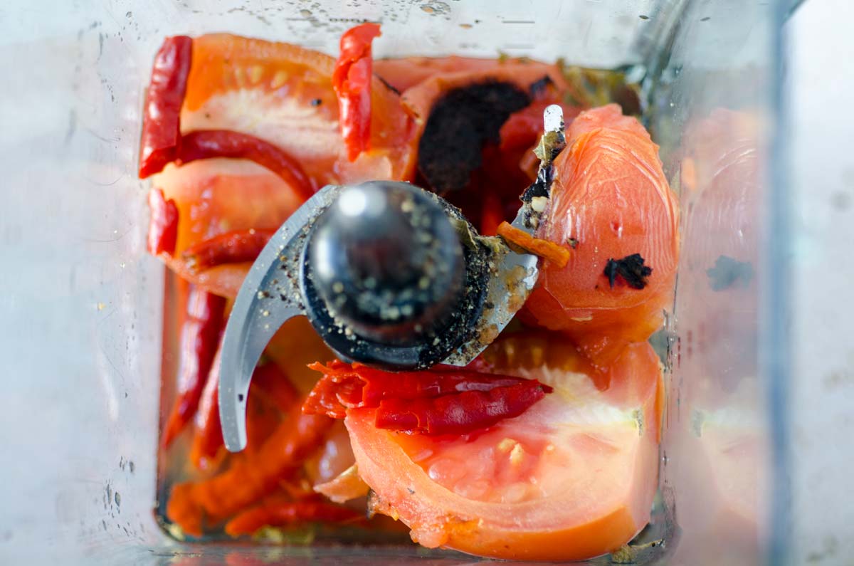 Top view of a blender jar containing peeled tomatoes and chili peppers.