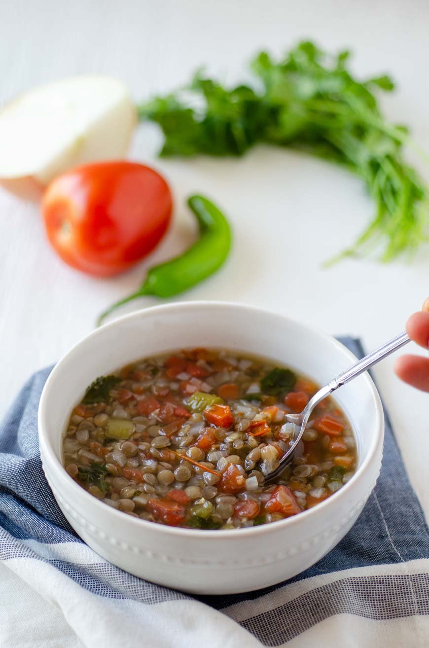 Mexican vegan lentil soup in a white bowl surrounded by tomato, cilantro and onion.