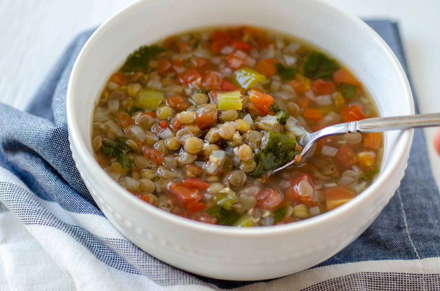 Mexican vegan lentil soup in a white bowl surrounded by tomato, cilantro and onion.