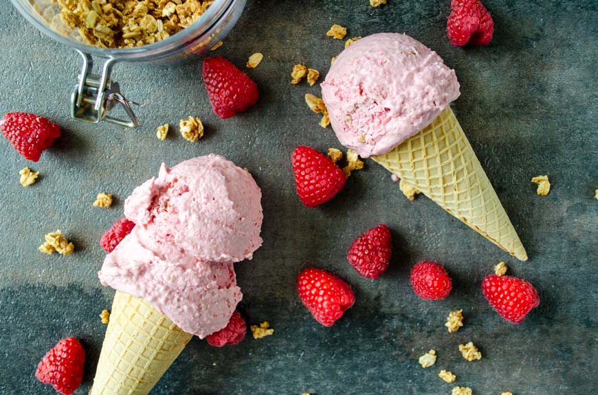 Two raspberry ice cream cones on top of a black background surrounded by oatmeal crumble and raspberries