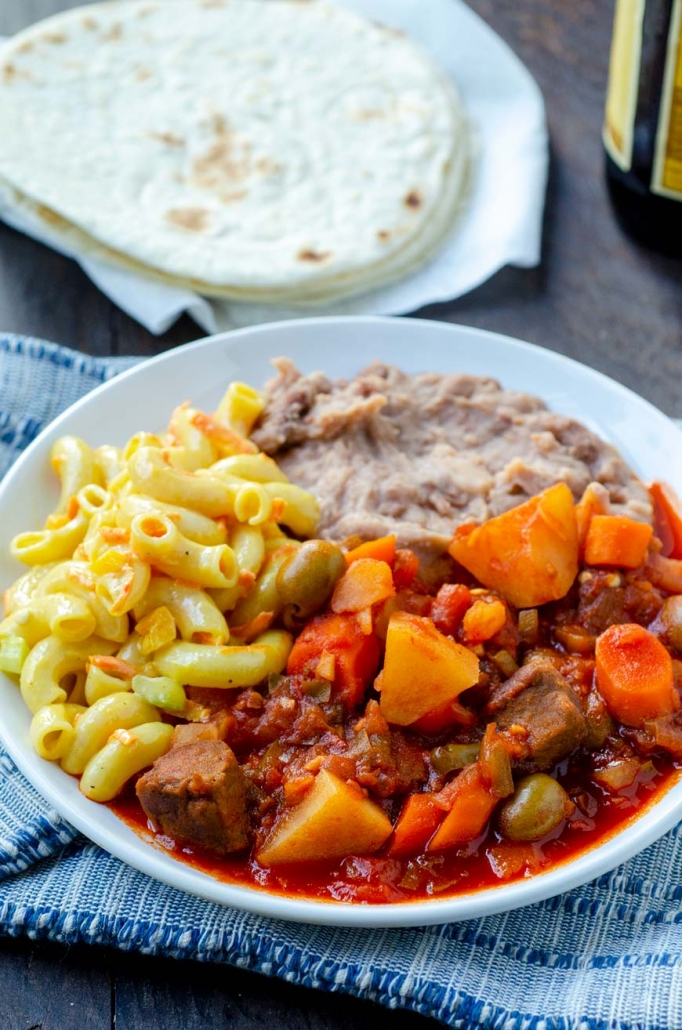 Vegan barbacoa sinaloense on a white plate with macaroni salad and refried beans, and flour tortillas.