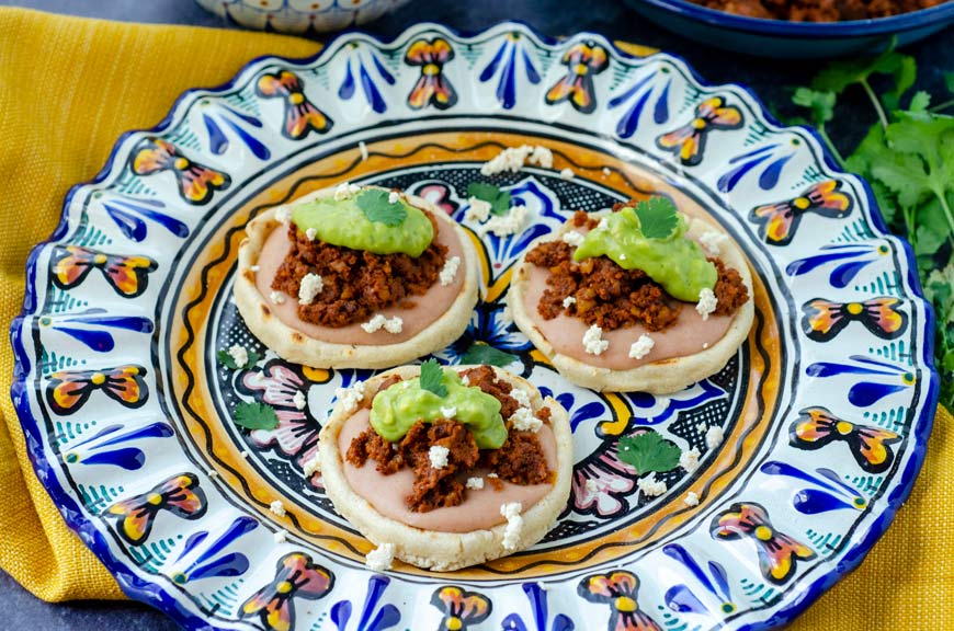 3 vegan sopes filled with beans, walnut meat, avocado salsa on a colorful talavera plate and yellow napkin underneath