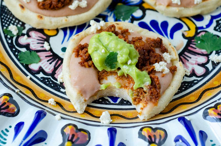 a sope filled with refried beans topped with walnut meat, cheese and avocado salsa with a bite taken out of it on a talavera plate