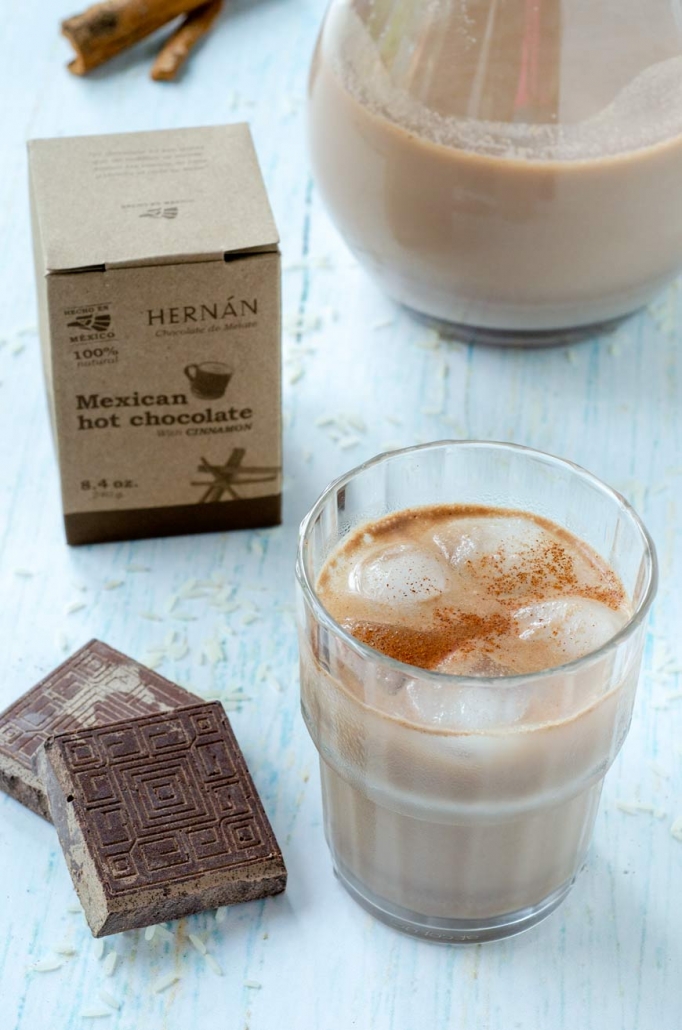a glass of chocolate horchata with 2 squares of chocolate beside it and the box of hernan chocolate behind it