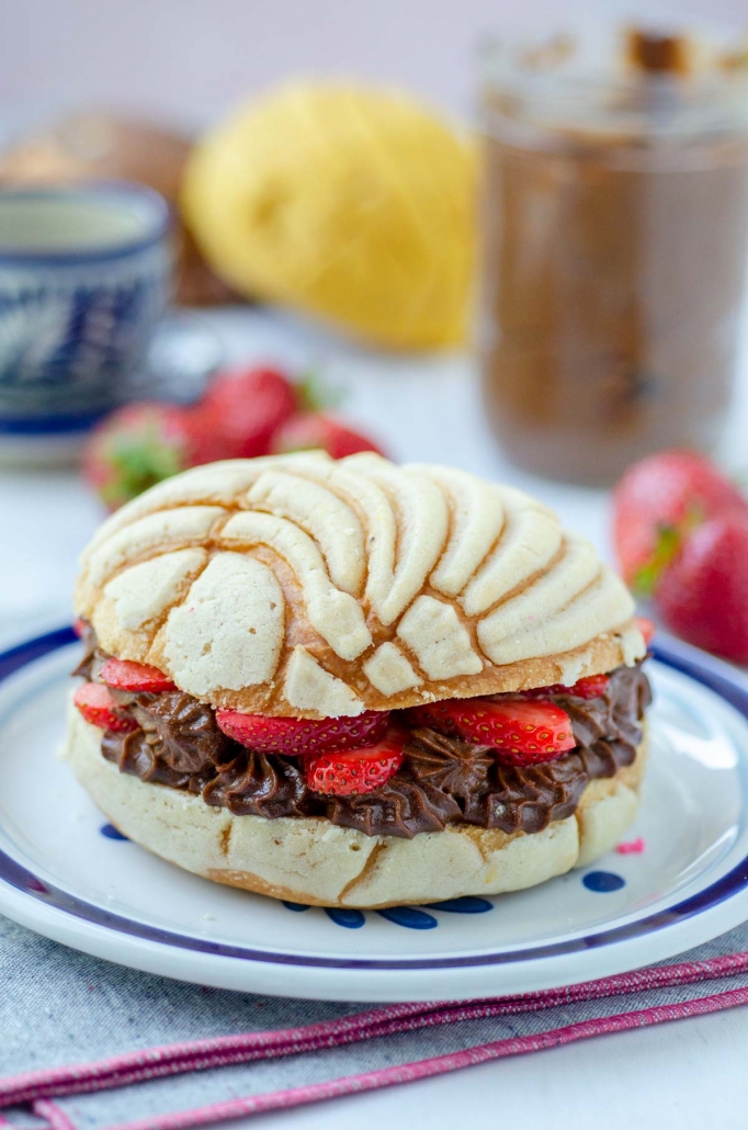 stuffed concha with vegan nutella and strawberries on a white and blue plate with strawberries in the background on a pink and gray napkin