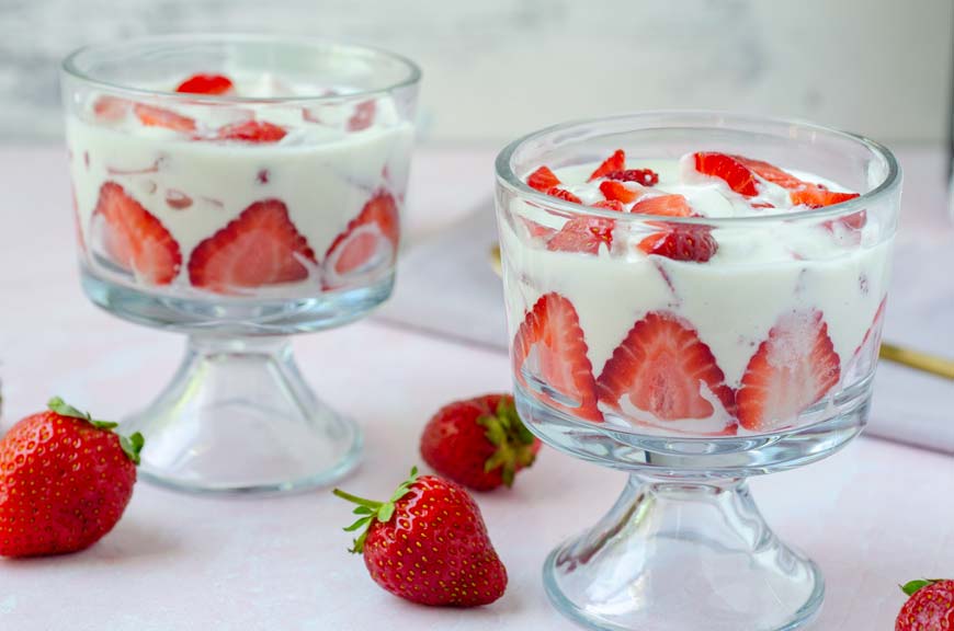 two glass cups filled with fresas con crema on a pink background surrounded by strawberries