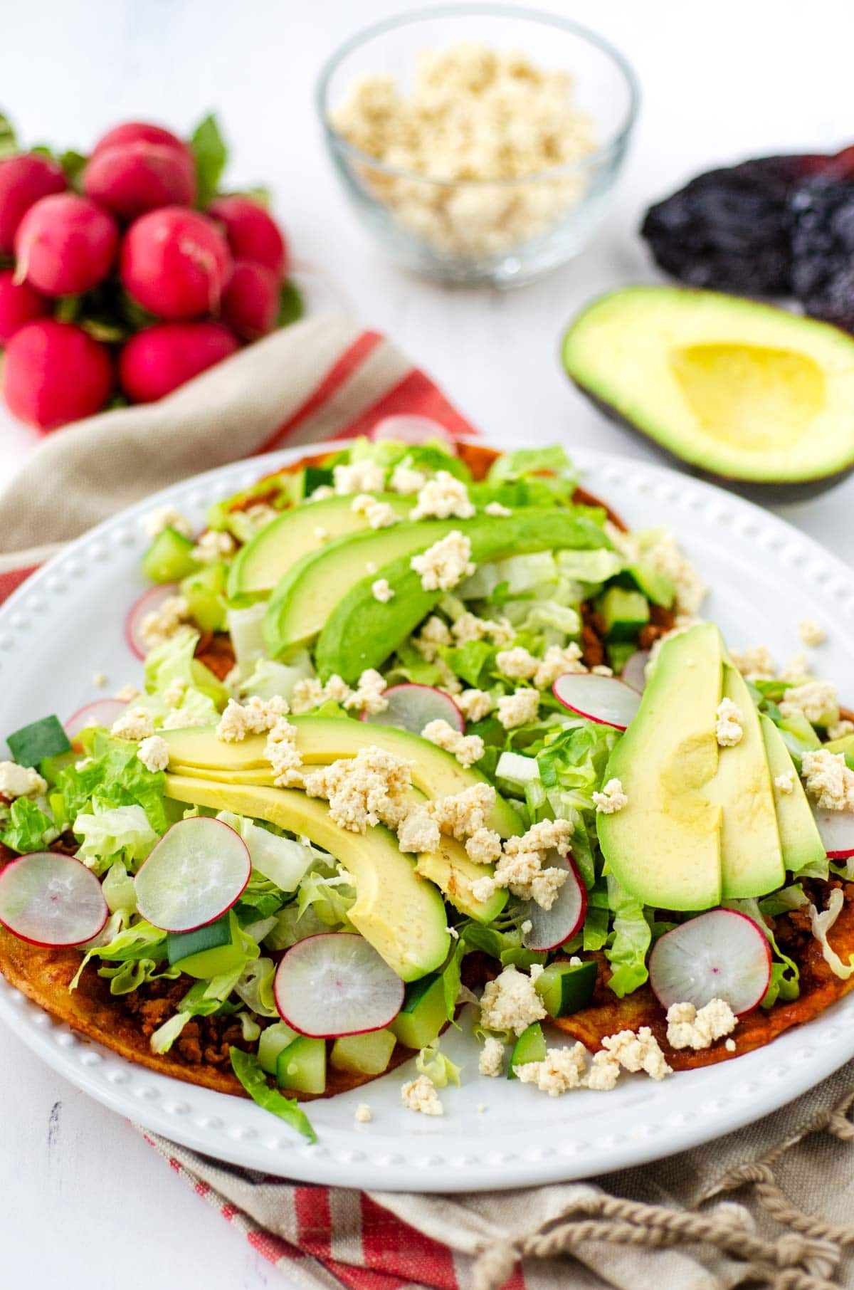 white plate with three tortillas topped with avocado, lettuce, radishes, queso fresco