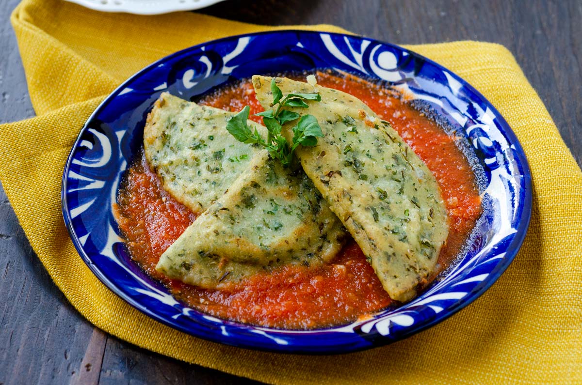 side view of three chaya empanadas on top of tomato sauce on blue plate