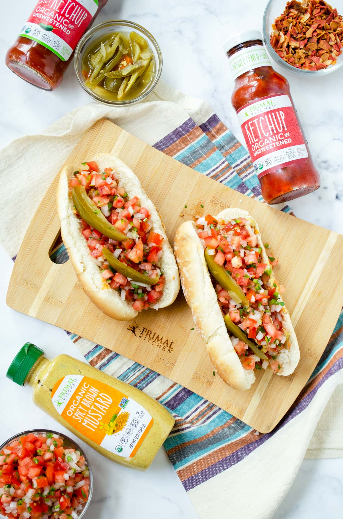 two Mexican hot dogs on a striped napkin with a marble background surrounded by ingredients