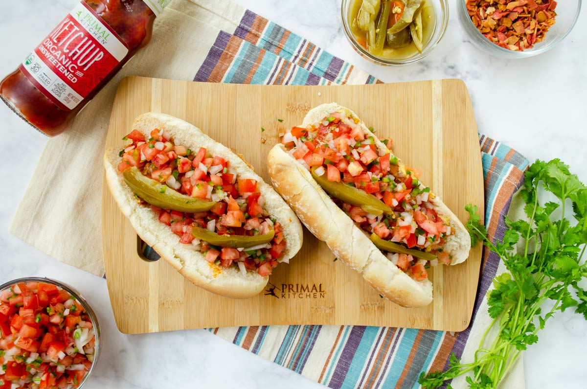 two hot dogs topped with pico de gallo on a colorful striped napkin surrounded by cilantro and a bottle of ketchup