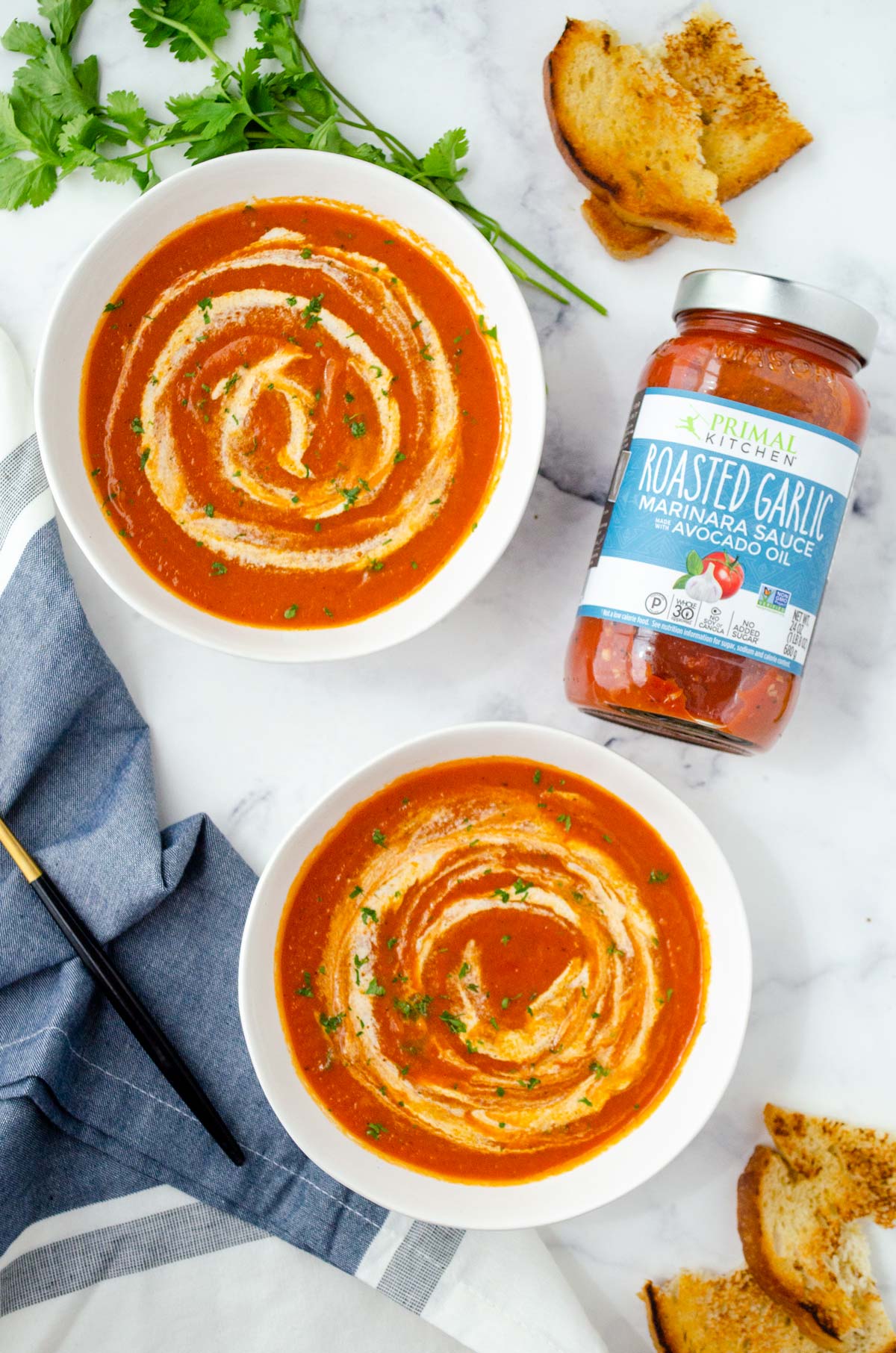 two bowls of vegan tomato soup with crema swirl by a jar of roasted garlic marinara