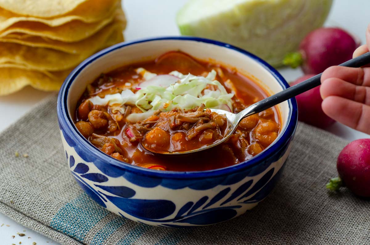 Vegan pozole in a white and blue talavera bowl on top of a linen napkin with a spoon digging in.