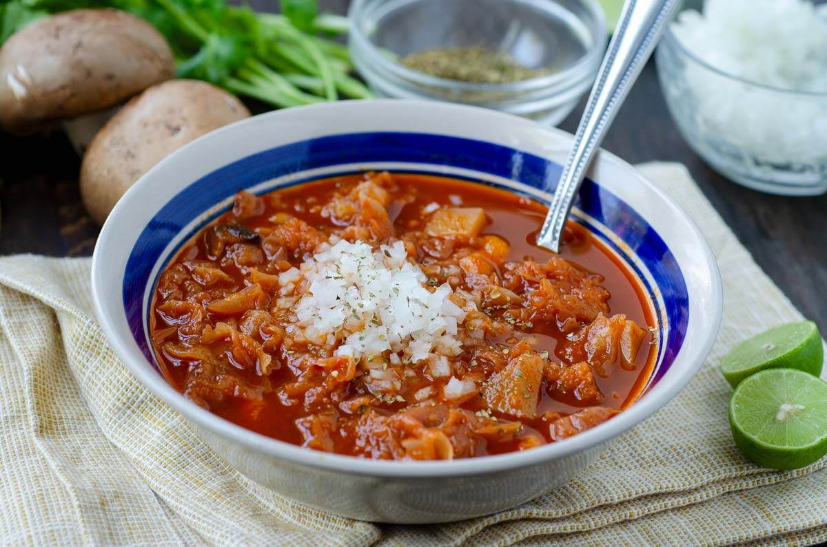 bowl of menudo surrounded by limes, mushrooms, oregano and onion