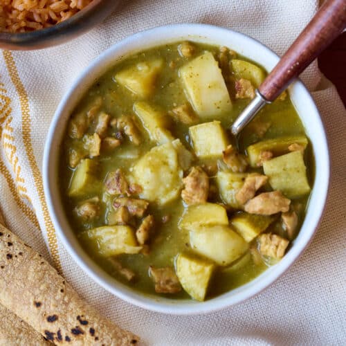 Bowl of chile verde on a white and yellow tablecloth, tortillas, and Mexican rice.