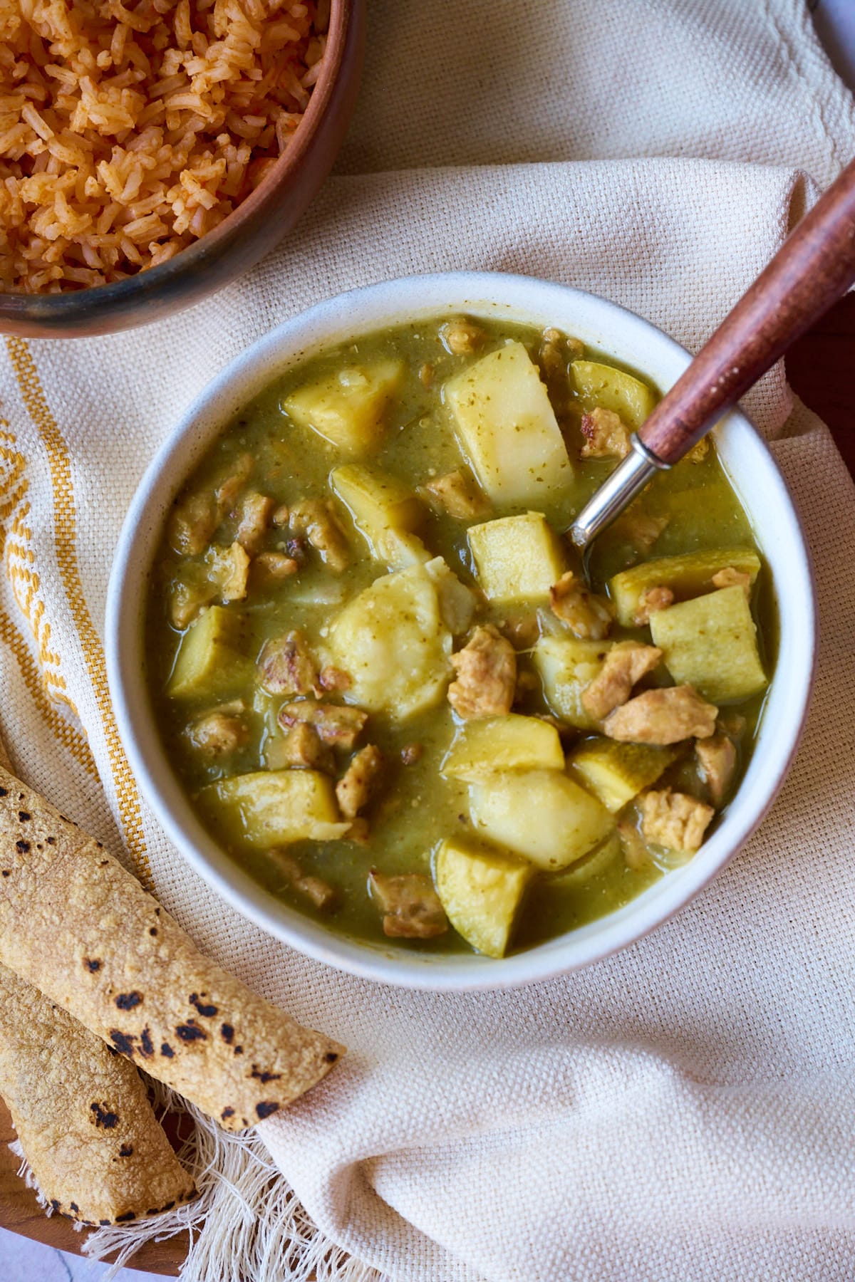 Bowl of chile verde on a white and yellow tablecloth, tortillas, and Mexican rice.