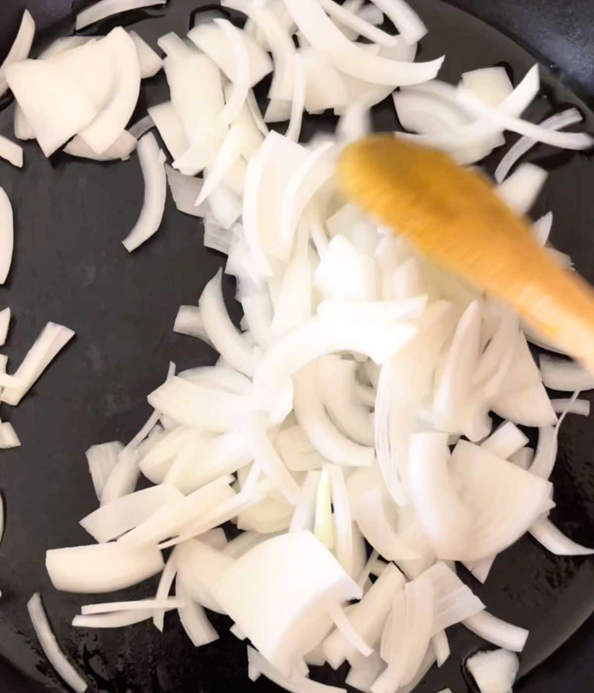 A wooden spoon stirring onions in a skillet.