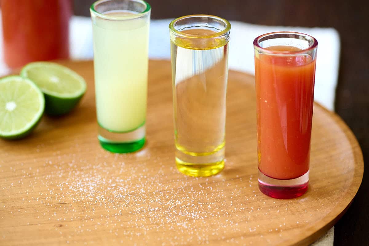 Three shot glasses, filled with sangrita, lime juice, and tequila on a wooden board with lime and salt.